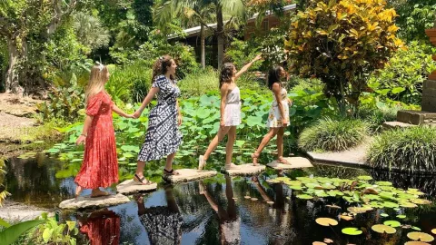 Four women cross a pond in a lush garden.