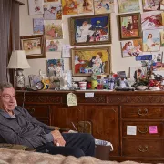 A man with tan skin and medium grey hair seated in front of a cluttered dresser and impressionist art Photo