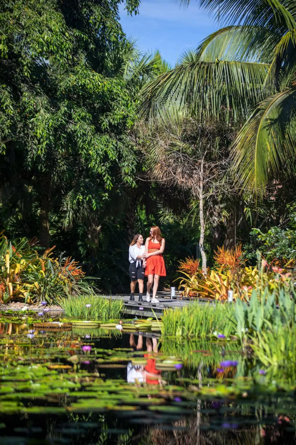 A queer couple explores a lush botanical garden with a pond.